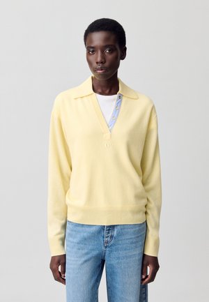 Maglione lavorato a maglia giallo chiaro con scollo a V, patta con bottoni blu e polsini a costine. Presenta un dettaglio del logo ricamato e una vestibilità rilassata.