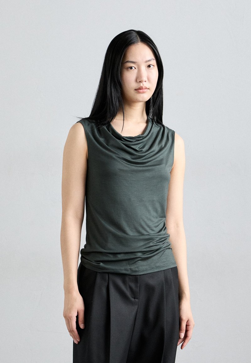 Dark green sleeveless top with cowl neckline and gathered texture, paired with loose black trousers. Simple, modern design.