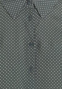 Green shirt with white polka dots, featuring a pointed collar and front button closure. Fabric appears smooth with a subtle shine.