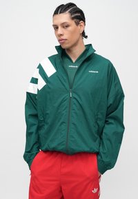 Osoba s vrkočmi v tmavozelenej športovej bunde Adidas s bielymi pruhmi a logom, v kombinácii s červenými nohavicami Adidas, stojaca pred bielym pozadím.