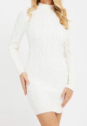 Kobieta ubrana w białą, długorękawową sukienkę sweterkową z warkoczowym splotem, z prążkowanym obrzeżem i dekoltem, pozująca z rękami przy biodrach.