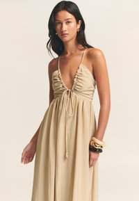 Vestido beige claro con escote pronunciado, tejido recogido y lazo ajustable. Textura suave y fluida. La modelo lleva pulseras en capas.