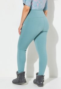 Persona che indossa leggings alto in vita color teal e stivali in camoscio grigio scuro per l'inverno, in piedi contro uno sfondo bianco.