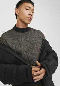Chaqueta negra acolchada con suéter de punto texturizado que presenta un cuello oscuro. El suéter tiene un sutil patrón moteado en gris y marrón.