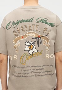 Πίσω μέρος μπεζ μπλουζάκι με πράσινο και κίτρινο κείμενο "Originals Studio UPSTATE, NY Queens 1990," ένα γραφικό λευκού λουλουδιού και χειρόγραφο κείμενο από κάτω.