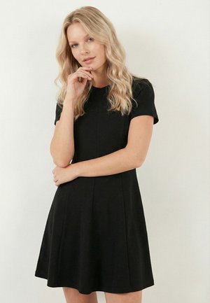 Femme blonde aux cheveux ondulés portant une robe noire à manches courtes, debout, une main touchant son menton et l'autre bras croisé.