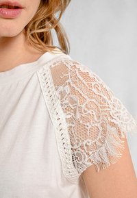 Top bianco con maniche a cappuccio in pizzo caratterizzate da intricati motivi floreali e dettagli a frange, abbinato a un corpo in tessuto morbido e semplice.