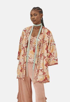 Bata estilo kimono con un patrón floral en tonos de beige y rojo, llevada sobre una blusa de color claro, combinada con pantalones suaves y texturizados en rosa.