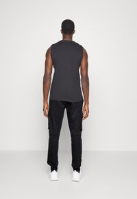 Svart ärmlös skjorta och mörka cargo-byxor med fickor. Modell vänder sig bort, bär vita sneakers. Slät textur, avslappnad design.