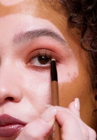 Close-up van een gezicht dat bruine eyeliner aanbrengt met een houten potlood. Het gezicht heeft een gladde huid, lichte make-up op de oogleden en een glanzende lippenstift.