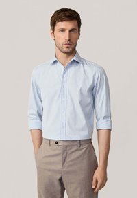 Camisa de botones a rayas azul claro con mangas largas remangadas, hecha de una tela suave. Combinada con pantalones beige a medida.