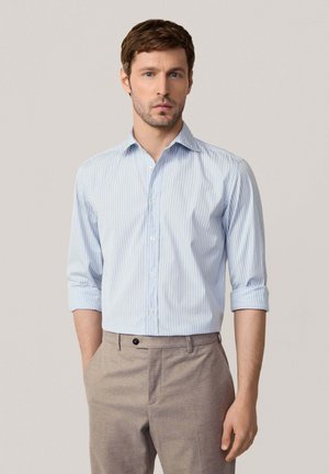 Camisa de botones a rayas azul claro con mangas largas remangadas, hecha de una tela suave. Combinada con pantalones beige a medida.