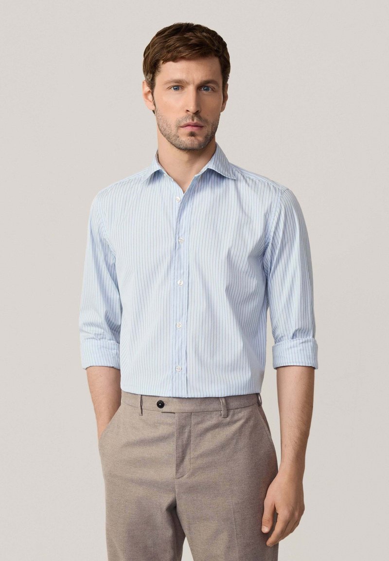 Camisa de botones a rayas azul claro con mangas largas remangadas, hecha de una tela suave. Combinada con pantalones beige a medida.