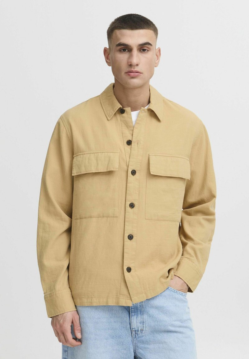 Chemise boutonnée beige clair avec deux poches poitrine, un col et des manches longues. Le tissu a une apparence texturée. Portée par-dessus un t-shirt blanc.