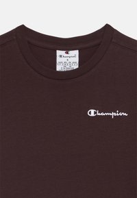 T-shirt in cotone marrone con collo tondo; presenta il logo "Champion" ricamato in bianco sul lato sinistro del petto; etichetta con taglia e istruzioni per la cura visibile.