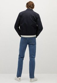 Mann trägt eine schwarze Jacke, blaue Jeans und weiße Sneakers, steht mit Blick auf eine schlichte hellgraue Wand und hat die Hände in den Taschen.