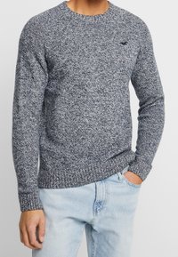 Man i långärmad, marinblå och vit melerad stickad tröja med ljusblå jeans, en hand i fickan mot en neutral bakgrund.