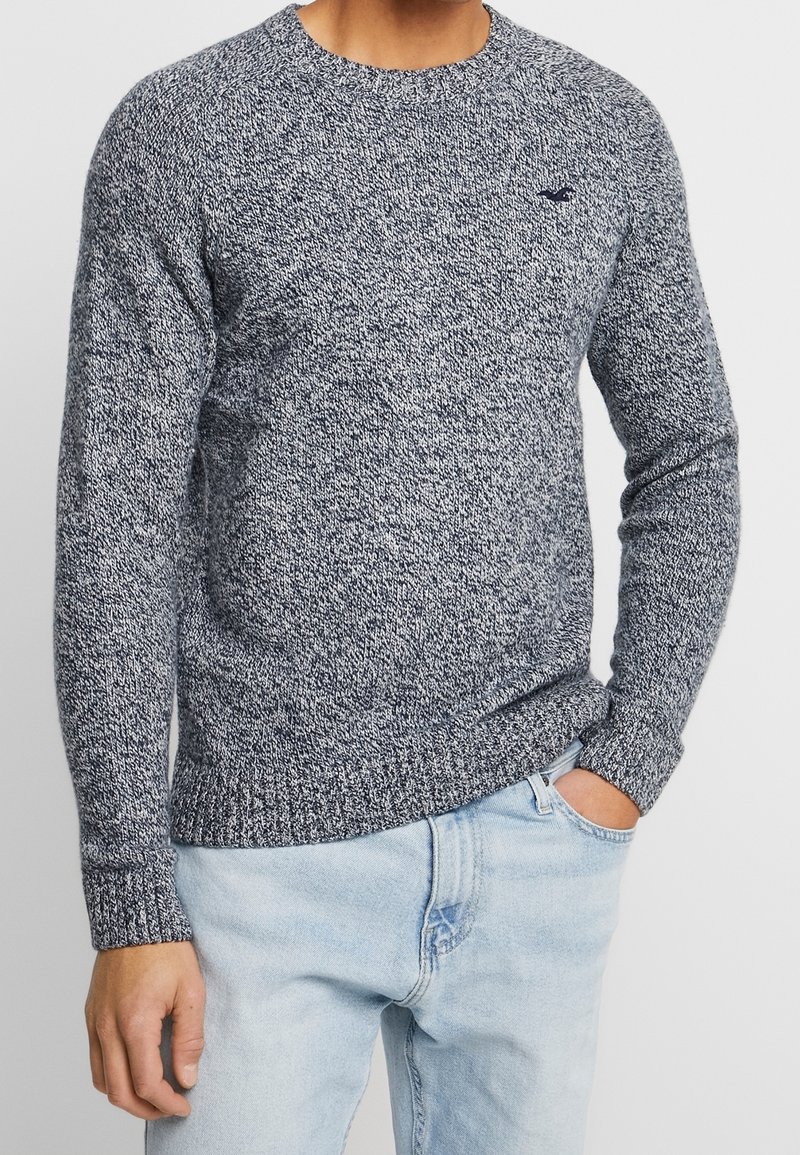 Man i långärmad, marinblå och vit melerad stickad tröja med ljusblå jeans, en hand i fickan mot en neutral bakgrund.