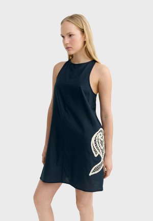 Frau trägt ärmelloses marineblaues Kleid mit hohem Ausschnitt und weißem Spitzenblumendetail an der linken Hüfte, steht vor einfarbigem Hintergrund.