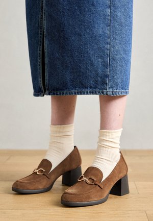 Brune ruskind sko med blokhæl, rundet tå og dekorativt metalbeslag. Brugt med cremefarvede ribstrikkede sokker og en blå denimnederdel.