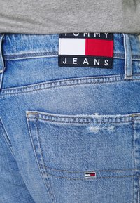 Světle modré džínové kalhoty s opotřebovanou strukturou, s tmavým štítkem na zadní straně s nápisem "TOMMY JEANS" a červenobílo-modrým logem.