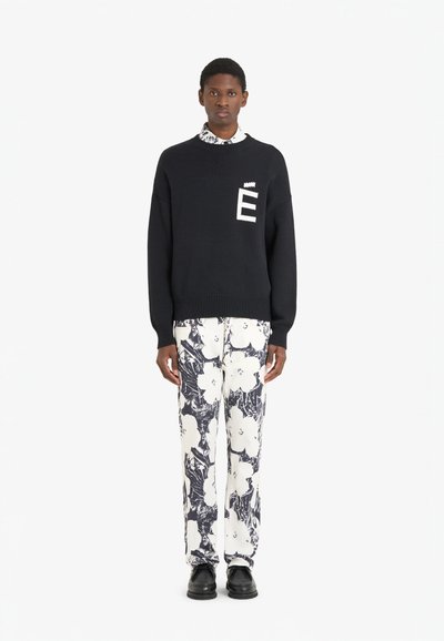 Pull en laine noir avec logo "É" blanc sur la poitrine, accompagné d'un pantalon blanc à motif floral noir et de chaussures noires.
