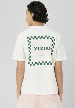 Personne aux cheveux bouclés portant un t-shirt blanc avec un carré à carreaux verts et le texte « MUCHO » dans le dos, les mains dans les poches, de dos.