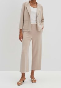 Beige sako s klopami, dvěma předními kapsami, spárované s odpovídajícími širokými kalhotami. Jednoduchý bílý top a tělové sandály dotvářejí vzhled.