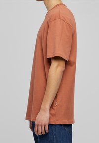 Hnědé tričko s krátkým rukávem, bavlněný materiál, kulatý výstřih, rovný střih, minimální detail postranního švu, nošeno s modrými džíny.