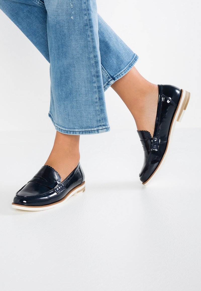 Personne portant un jean bleu clair et des mocassins penny noirs brillants avec des semelles blanches, debout sur une surface blanche.