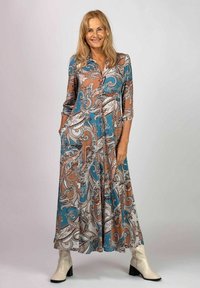 Lång, paisley-mönstrad klänning i teal, brunt och vitt; har krage, trekvartslånga ärmar och fickor. Bärs med krämfärgade klackstövlar.