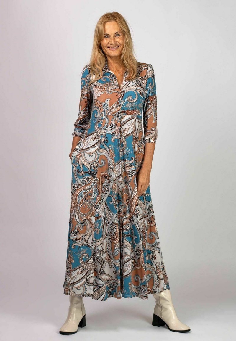 Lång, paisley-mönstrad klänning i teal, brunt och vitt; har krage, trekvartslånga ärmar och fickor. Bärs med krämfärgade klackstövlar.