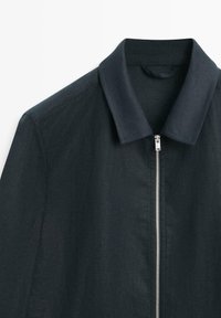 Veste bleu marine foncé avec un col rabattable et une fermeture éclair argentée centrée, montrée de la poitrine supérieure au col.