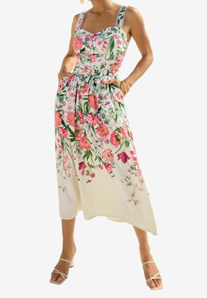 Femme portant une robe midi blanche sans manches avec un imprimé floral rose et vert, des poches, et des sandales à talons blanches à lanières.
