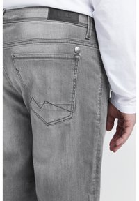 Graue Denim-Jeans mit strukturiertem Finish, das ein einzigartiges gestepptes Taschen-Design und ein schwarzes Lederetikett am Bund aufweist.