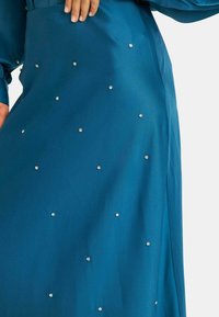 Vestido de satén verde azulado con una textura suave, adornado con tachuelas metálicas dispersas en la tela para darle detalle. Mangas largas y diseño fluido.
