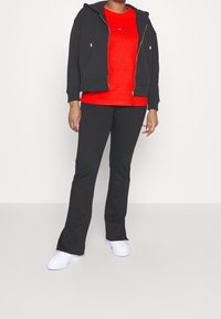 Sort hættejakke med lynlås, rød t-shirt med tekst og sorte flared bukser. Hvide sneakers fuldender outfittet. Glat struktur hele vejen igennem.