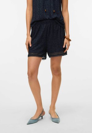 Vrouw draagt marineblauwe kanten korte broek en bijpassende top, blauwe puntige slingback hakken en een houten armband om haar pols.