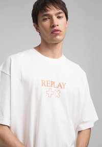 T-shirt en coton blanc à manches courtes, présentant un logo orange "REPLAY" et des symboles "+" centrés sur la poitrine. Design simple, coupe décontractée.