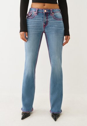 Femme portant un jean évasé bleu clair avec des coutures contrastantes rose vif, associé à des chaussures noires à bouts pointus et un crop top noir à manches longues.