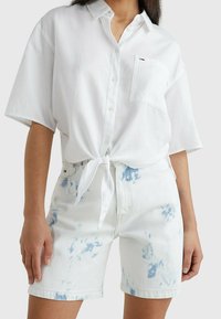 Camicia bianca con maniche corte, legata in vita; abbinata a un paio di pantaloni corti di jeans chiari con motivi tie-dye blu e tasche.