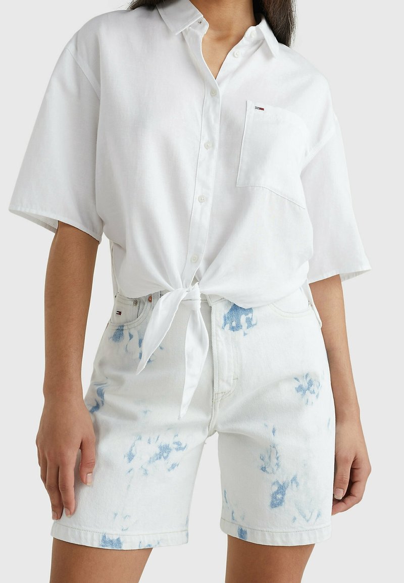 Camicia bianca con maniche corte, legata in vita; abbinata a un paio di pantaloni corti di jeans chiari con motivi tie-dye blu e tasche.