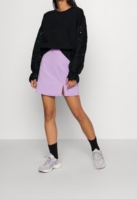 Maglione nero a maniche lunghe con maniche testurizzate, abbinato a una mini gonna lilla con uno spacco frontale. Indossato con sneakers grigie e calze nere.
