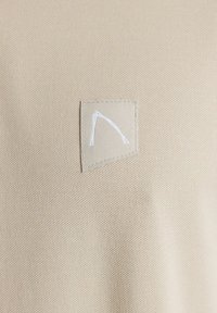 Tessuto beige con texture e un'etichetta quadrata con un design bianco. Il materiale presenta un motivo fine e traspirante.