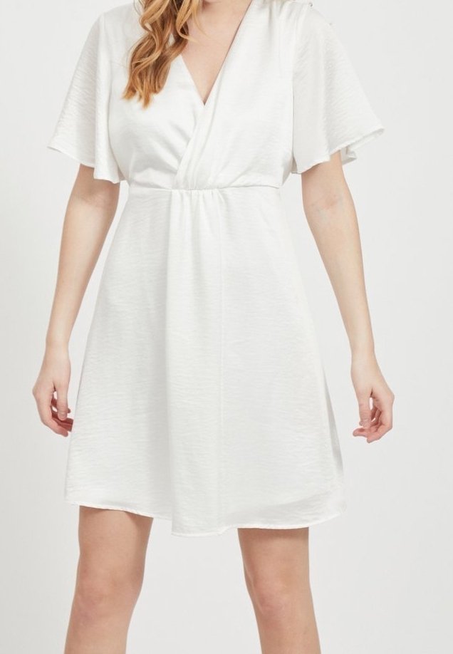 Femme portant une courte robe blanche avec des manches flottantes et un col en V, debout devant un fond clair uni.