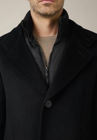 Czarny płaszcz z mieszanki wełny z klapami, z zamkiem błyskawicznym w czarnej wewnętrznej kurtce i jasnobeżowym swetrem pod spodem.