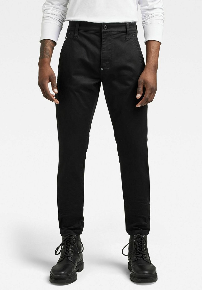 Homme portant un pantalon noir slim, des bottes noires à lacets et une chemise blanche à manches longues, debout devant un fond blanc uni.