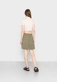 Mulher de costas, vestindo uma camisa polo curta bege, uma saia em camadas verde-oliva e sandálias pretas, sobre um fundo branco simples.