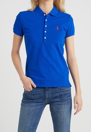 Femme portant un polo bleu à manches courtes avec un logo rouge brodé et un jean bleu, debout avec une main dans la poche.