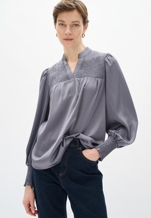Blusa grigia con vestibilità comoda, parte superiore arricciata, maniche lunghe e scollatura a V. Abbinata a jeans in denim scuro. Tessuto morbido e lucentezza delicata.